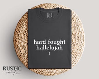 May include: A dark gray t-shirt with the words "hard fought hallelujah" and a cross printed in white. The shirt is displayed on a round, woven wicker placemat.