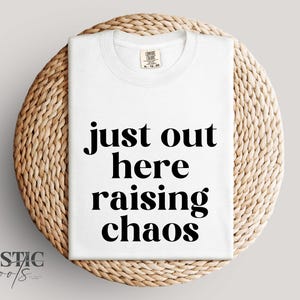 May include: White t-shirt with the text "just out here raising chaos" in black, displayed on a round woven placemat. The shirt has a crew neck and short sleeves. The brand name "Rustic Roots" is in the lower left corner.