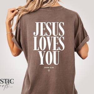 May include: A brown t-shirt with the text "JESUS LOVES YOU" in large white letters on the back. Below the text is "JOHN 3:16" and a small cross. The shirt has rolled-up sleeves.