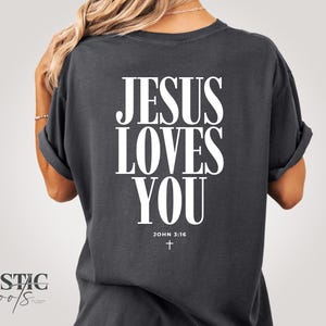 May include: Dark gray t-shirt with the text "JESUS LOVES YOU" in large white letters. Below the text is "JOHN 3:16" and a small cross. The shirt has rolled-up sleeves.
