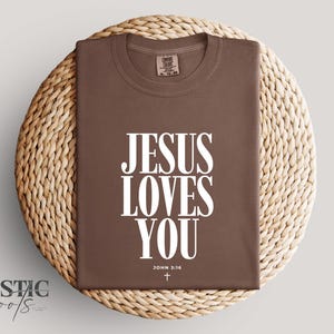 May include: A brown t-shirt with the text "JESUS LOVES YOU" in large white letters. Below the text is "JOHN 3:16" and a small cross. The shirt is displayed on a woven, circular surface.