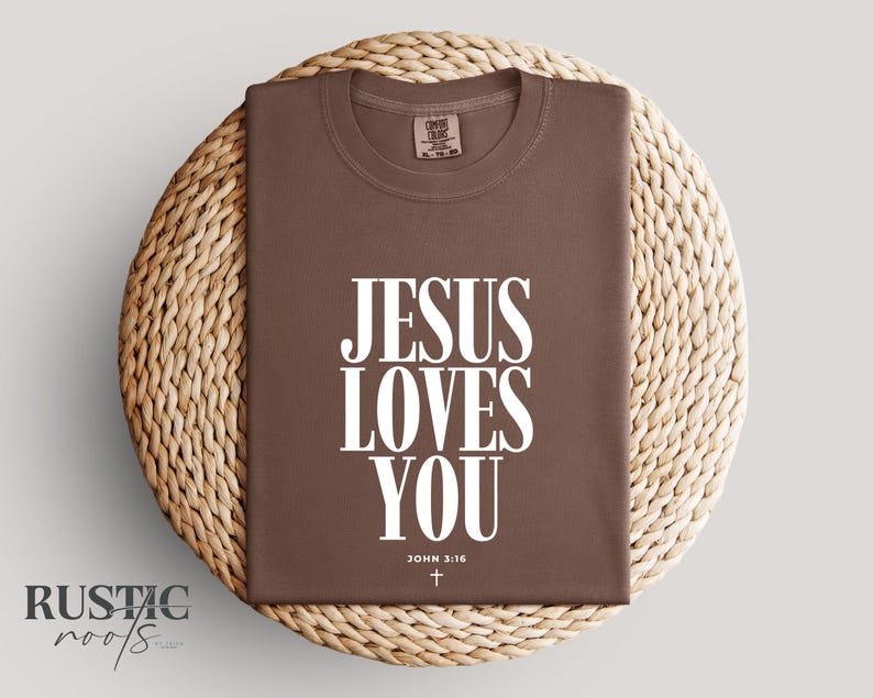 May include: A brown t-shirt with the text "JESUS LOVES YOU" in large white letters. Below the text is "JOHN 3:16" and a small cross. The shirt is displayed on a woven, circular surface.