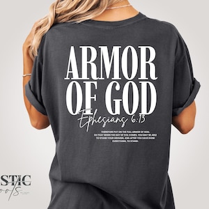 May include: Dark grey t-shirt featuring the text "ARMOR OF GOD" in large white letters. Below the text is "Ephesians 6:13" and a Bible verse. The shirt has rolled-up sleeves and a relaxed fit.