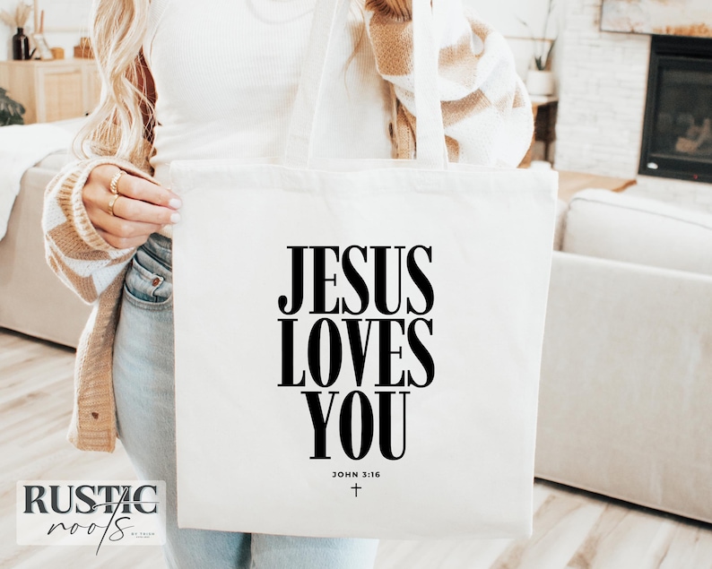 May include: A white tote bag with the text "JESUS LOVES YOU" in large black letters. The text "JOHN 3:16" and a small cross are below the main text. The bag has white handles.