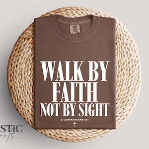 Peut inclure: Un t-shirt marron avec l'inscription blanche "WALK BY FAITH NOT BY SIGHT", la référence biblique 2 Corinthiens 5:7 et une petite croix. Le t-shirt est présenté sur un tapis rond en osier.