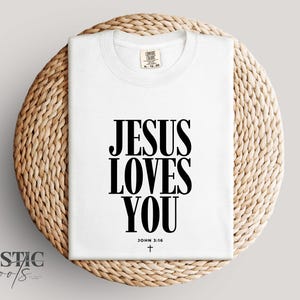 May include: A white t-shirt with the text "JESUS LOVES YOU" in bold black letters. The text "JOHN 3:16" and a small cross are below the main text. The shirt is folded and placed on a woven, circular mat.