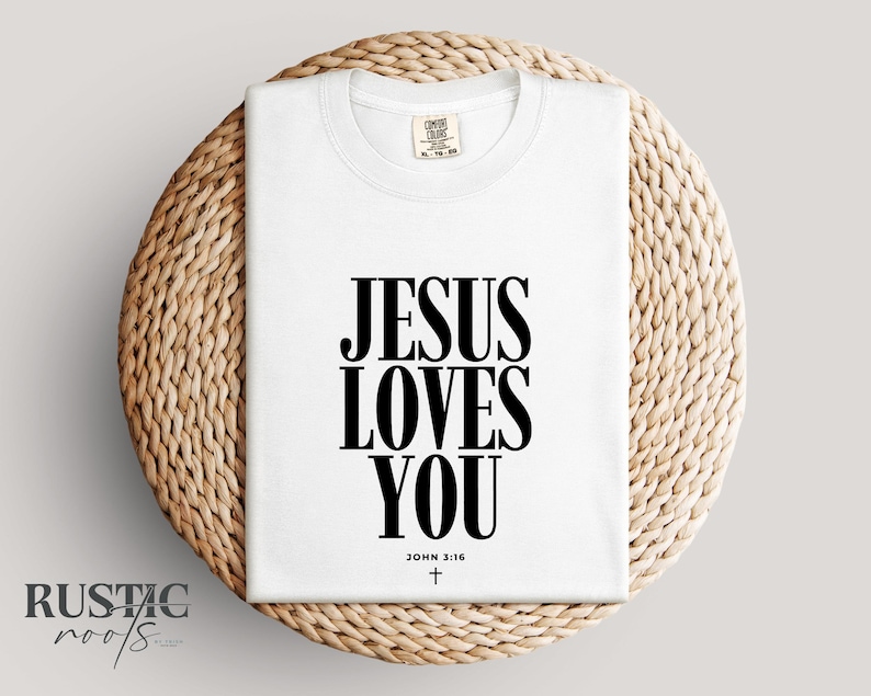 May include: A white t-shirt with the text "JESUS LOVES YOU" in bold black letters. The text "JOHN 3:16" and a small cross are below the main text. The shirt is folded and placed on a woven, circular mat.