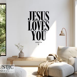 May include: A living room scene with a white wall featuring the text "JESUS LOVES YOU" in large black letters. Below the text is "JOHN 3:16" and a cross. The room includes a beige armchair, ottoman, and wooden bench.