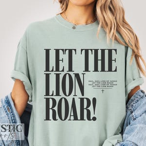 Pode incluir: Camiseta verde claro com a frase "LET THE LION ROAR!" em letras pretas grandes. Abaixo, o texto "HAIL, HAIL LION OF JUDAN LET THE LION ROAR!" e uma pequena cruz. Uma peça de roupa casual.