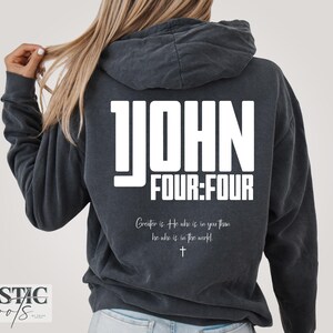 Op de afbeelding: Een antracietgrijze hoodie met witte tekst op de achterkant die 1 JOHN FOUR:FOUR leest, gevolgd door de cursieve tekst Greater is He who is in you than he who is in the world, en een klein kruissymbool in het midden eronder.