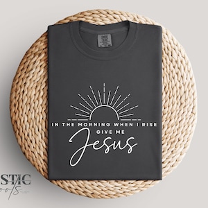 May include: Dark gray t-shirt with white text and a sunburst design. The text reads "IN THE MORNING WHEN I RISE GIVE ME Jesus". The shirt is displayed on a woven, circular surface.