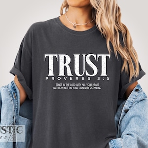 May include: Dark gray t-shirt with the word "TRUST" in large white letters. Below, it reads "PROVERBS 3:5" and a longer quote. The shirt is part of the "Rustic Roots" collection.