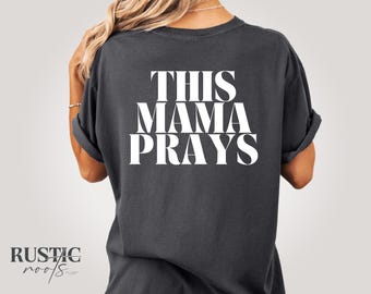 May include: Dark gray t-shirt with the text "THIS MAMA PRAYS" in large white letters on the back. The shirt has rolled-up sleeves and a relaxed fit. The Rustic Roots logo is visible in the bottom left corner.