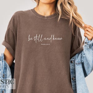 May include: A brown t-shirt with the text "be still and know" in a cursive font. The text "Psalms 46:10" is below the main text. The shirt is worn with a denim jacket.
