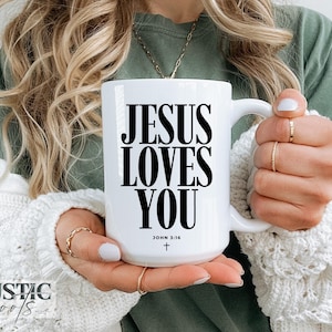 May include: White ceramic mug with the text "JESUS LOVES YOU" in black, along with "JOHN 3:16" and a cross. The mug is held by a person wearing a white sweater and gold rings. The mug has a large handle.
