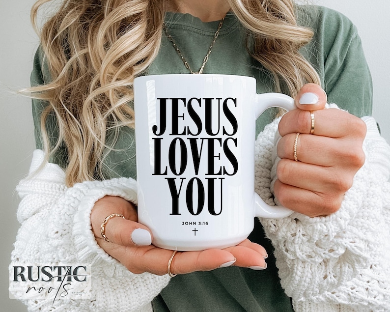 May include: White ceramic mug with the text "JESUS LOVES YOU" in black, along with "JOHN 3:16" and a cross. The mug is held by a person wearing a white sweater and gold rings. The mug has a large handle.