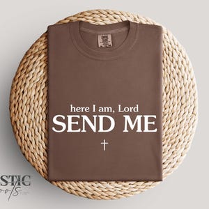 Op de afbeelding: Een bruin t-shirt met de tekst "here I am, Lord SEND ME" in het wit, en een wit kruis. Het shirt wordt getoond op een ronde, geweven placemat van riet. De merknaam "Rustic Roots" staat linksonder.