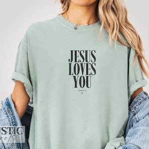 May include: A light green t-shirt with the text "JESUS LOVES YOU" in black, with "JOHN 3:16" and a cross below. The shirt has rolled-up sleeves and is made of a soft material.
