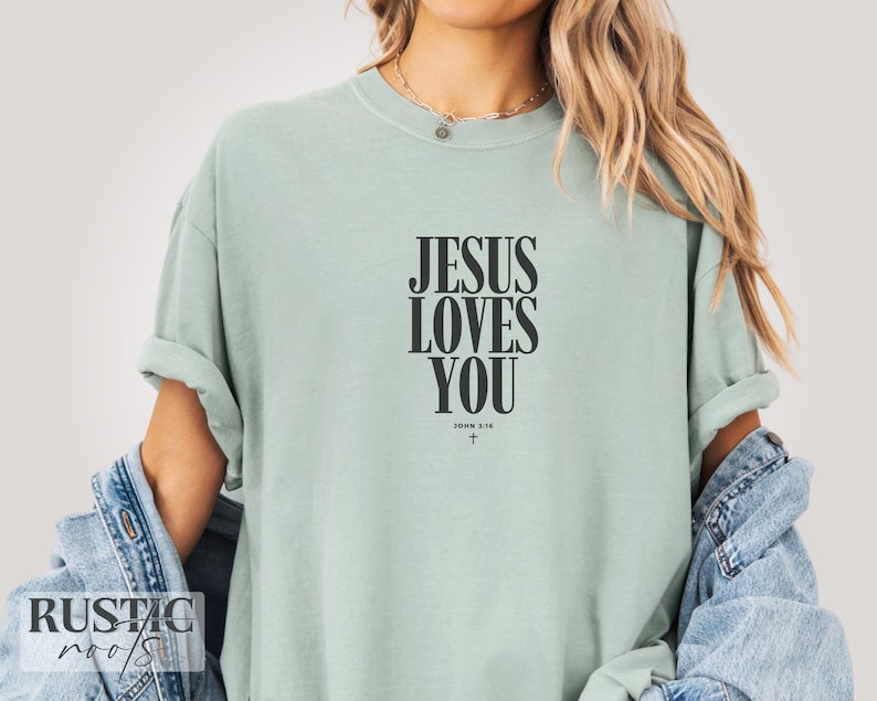 May include: A light green t-shirt with the text "JESUS LOVES YOU" in black, with "JOHN 3:16" and a cross below. The shirt has rolled-up sleeves and is made of a soft material.