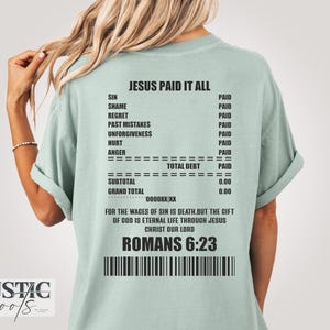 May include: A pale green t-shirt with the words "JESUS PAID IT ALL" and a list of sins marked as "PAID". The shirt also features the Bible verse Romans 6:23 and a barcode design.