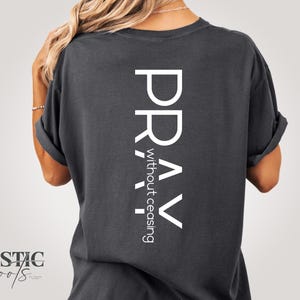 May include: Dark gray t-shirt with the words "PRAY without ceasing" printed vertically in white. The shirt has rolled-up sleeves and a relaxed fit. The brand logo "Rustic Roots" is at the bottom left.