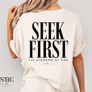 May include: Cream-colored t-shirt with the words "SEEK FIRST" in bold black letters on the back. Below the text are the words "THE KINGDOM OF GOD" and "Matthew 6:33". The shirt has rolled-up sleeves.