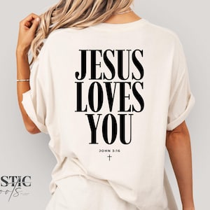 May include: Cream-colored t-shirt with the message "JESUS LOVES YOU" in bold black letters on the back. The text "JOHN 3:16" and a small cross are below the main text. The shirt has rolled-up sleeves.