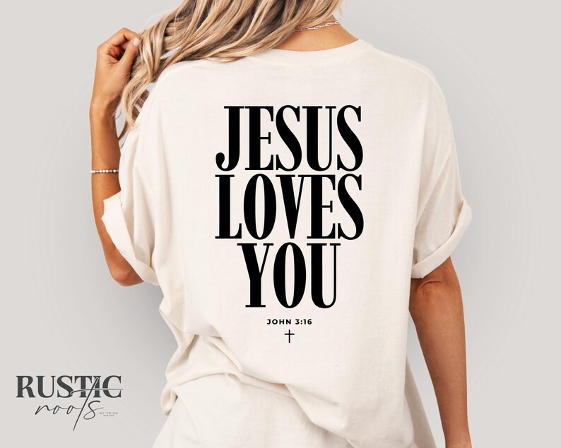 May include: Cream-colored t-shirt with the message "JESUS LOVES YOU" in bold black letters on the back. The text "JOHN 3:16" and a small cross are below the main text. The shirt has rolled-up sleeves.
