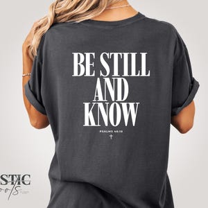 Peut inclure: T-shirt gris foncé avec l'inscription blanche "BE STILL AND KNOW", ainsi que "PSALMS 46:10" et une petite croix. Le t-shirt a des manches retroussées et une coupe décontractée.