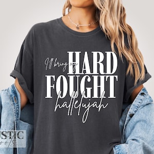 May include: Dark gray t-shirt with the text "I'll bring my HARD FOUGHT hallelujah" in white. The shirt has rolled-up sleeves and a relaxed fit. The brand name "RUSTIC roots" is visible at the bottom left.