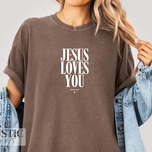May include: A brown t-shirt with the text "JESUS LOVES YOU" in white, with "JOHN 3:16" and a small cross underneath. The shirt has rolled-up sleeves and is worn with a denim jacket.