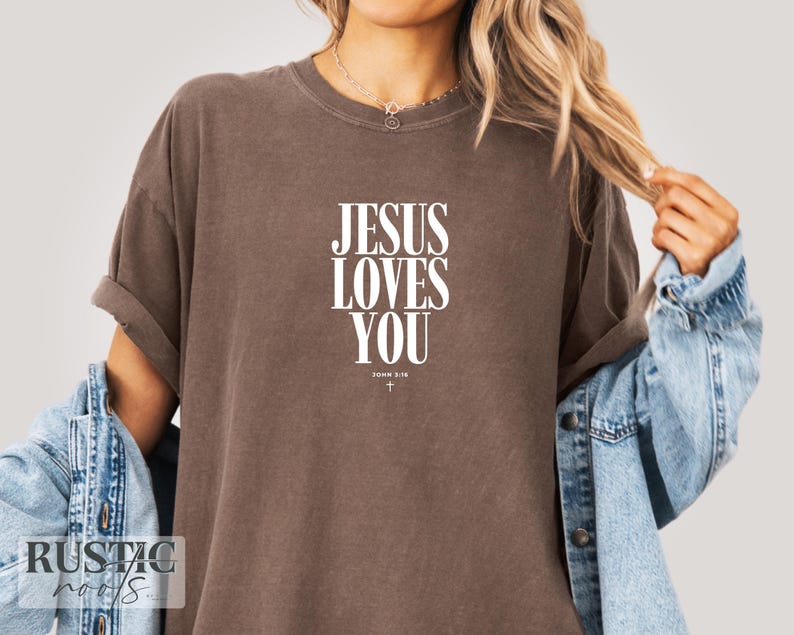 May include: A brown t-shirt with the text "JESUS LOVES YOU" in white, with "JOHN 3:16" and a small cross underneath. The shirt has rolled-up sleeves and is worn with a denim jacket.