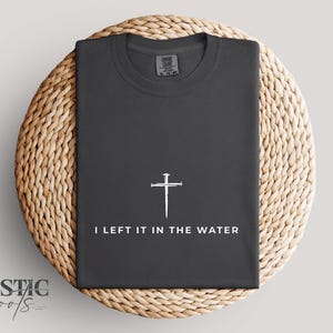 May include: Dark gray t-shirt with a white cross and the text "I LEFT IT IN THE WATER." The shirt is folded and placed on a round, woven, light brown surface. The brand name "RUSTIC roots" is in the lower left corner.