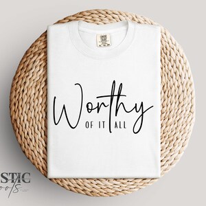 May include: A white t-shirt with the words "Worthy of it All" in black script. The shirt is folded and placed on a round, woven, light brown placemat. The brand name "Rustic Roots" is in the lower left corner.