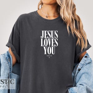 May include: Dark gray t-shirt with the text "JESUS LOVES YOU" in white, with "JOHN 3:16" and a cross below. The shirt has rolled-up sleeves and is worn with a denim jacket.