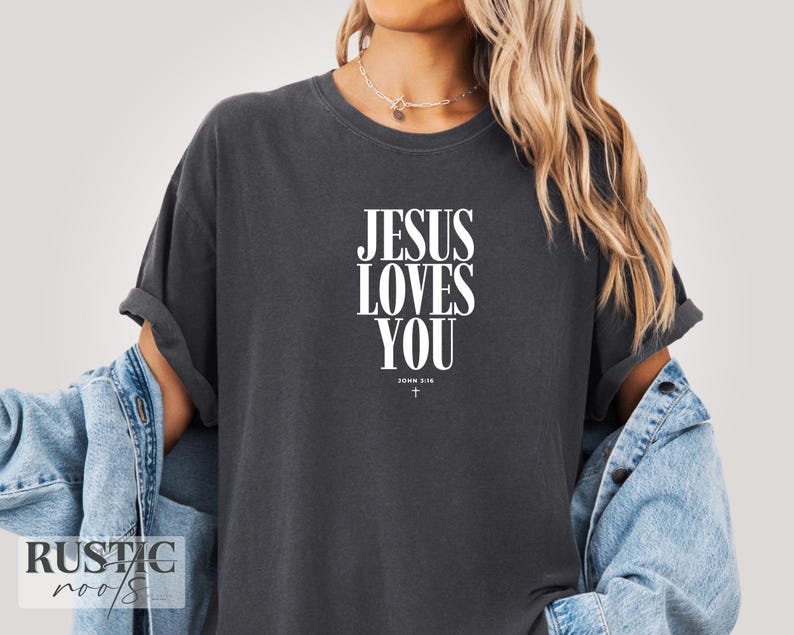 May include: Dark gray t-shirt with the text "JESUS LOVES YOU" in white, with "JOHN 3:16" and a cross below. The shirt has rolled-up sleeves and is worn with a denim jacket.