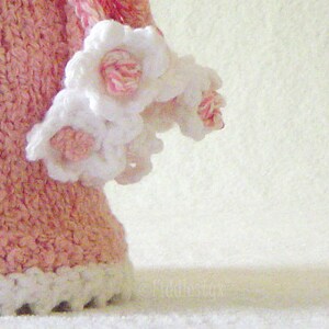 Puede incluir: Un vestido de crochet rosa con flores de crochet blancas alrededor del dobladillo inferior.