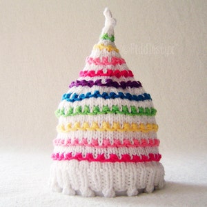 May include: White knitted baby hat with a rainbow stripe pattern. The hat has a pointed top and a white pom-pom.