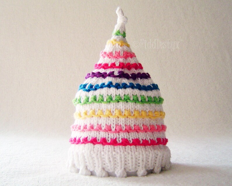 May include: White knitted baby hat with a rainbow stripe pattern. The hat has a pointed top and a white pom-pom.