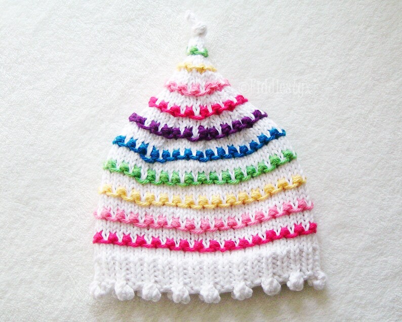 May include: White knitted hat with a rainbow pattern of colorful yarn. The hat has a pointed top and a scalloped brim.