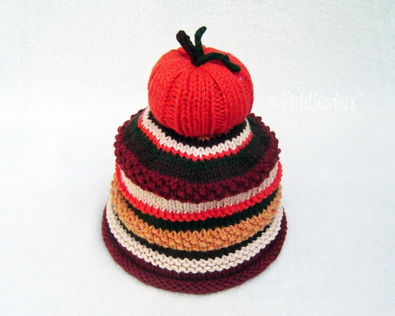 Puede incluir: Un gorro de punto con un dise&ntilde;o en capas en tonos de rojo, naranja, marr&oacute;n y blanco. La capa superior tiene forma de tomate.