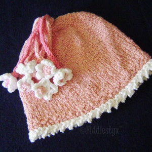 Puede incluir: Un gorro de punto rosa con ribete blanco de ganchillo y adornos de flores blancas.