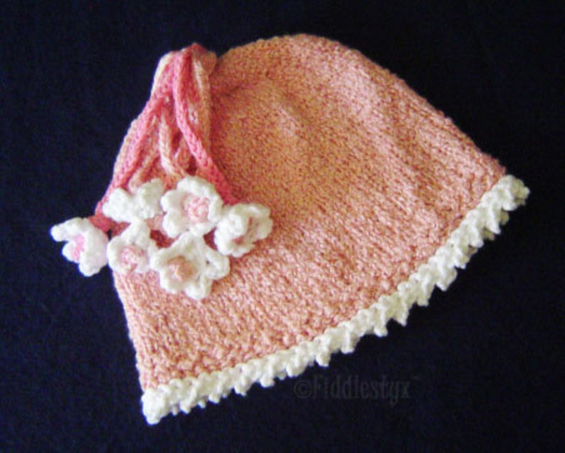 Puede incluir: Un gorro de punto rosa con ribete blanco de ganchillo y adornos de flores blancas.
