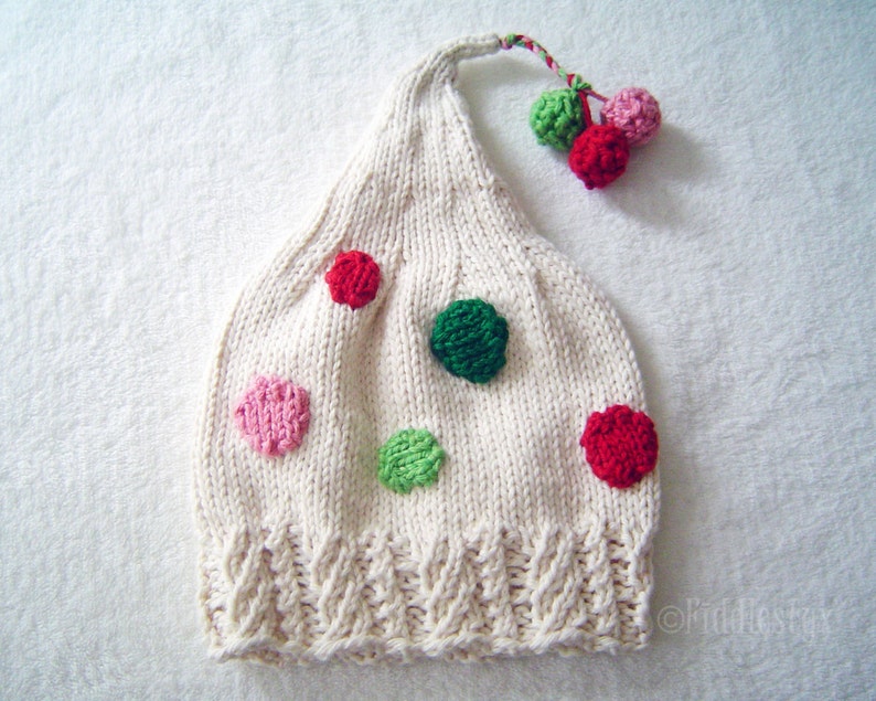 May include: A white knitted hat with a pointed top and a pom-pom on the end. The hat is decorated with colourful polka dots in pink, green, and red.