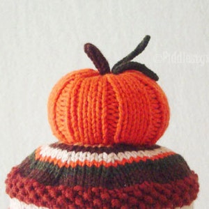 Puede incluir: Un gorro de punto de calabaza con rayas marrones, blancas y rojas. El gorro est&aacute; coronado con una calabaza naranja de punto.