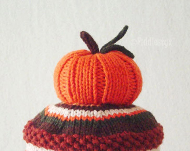Puede incluir: Un gorro de punto de calabaza con rayas marrones, blancas y rojas. El gorro est&aacute; coronado con una calabaza naranja de punto.