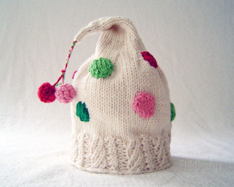 May include: White knitted hat with a pointed top and a long tail. The hat is decorated with colourful polka dots in shades of pink, green, and red.