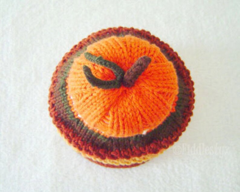 Puede incluir: Un gorro de punto de calabaza con rayas naranjas, marrones y verdes. El gorro est&aacute; hecho de hilo y tiene una superficie texturizada.