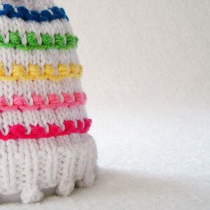 May include: White crocheted hat with a colorful rainbow stripe pattern. The hat has a pom pom on top and a fringe around the bottom.