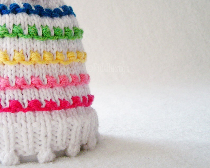 May include: White crocheted hat with a colorful rainbow stripe pattern. The hat has a pom pom on top and a fringe around the bottom.